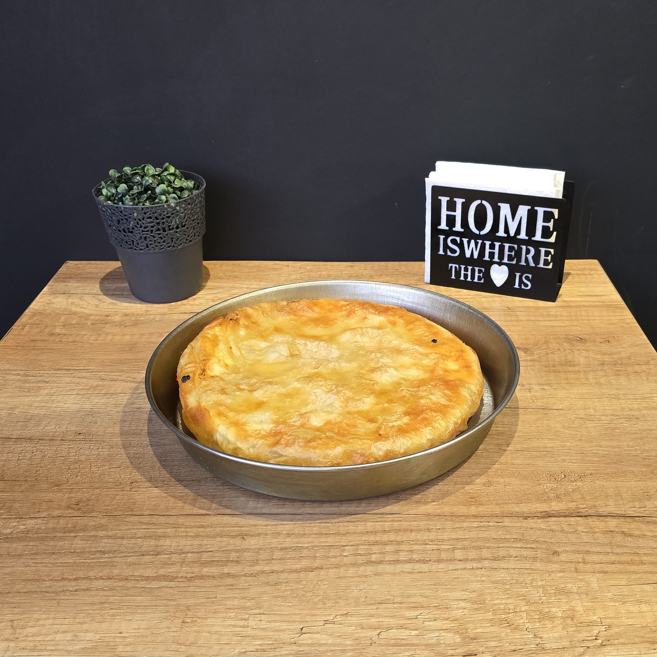 Golden-brown baked pie in a round metal pan on a wooden table with a small potted plant and a black napkin holder that reads 'HOME IS WHERE THE ♥ IS'.