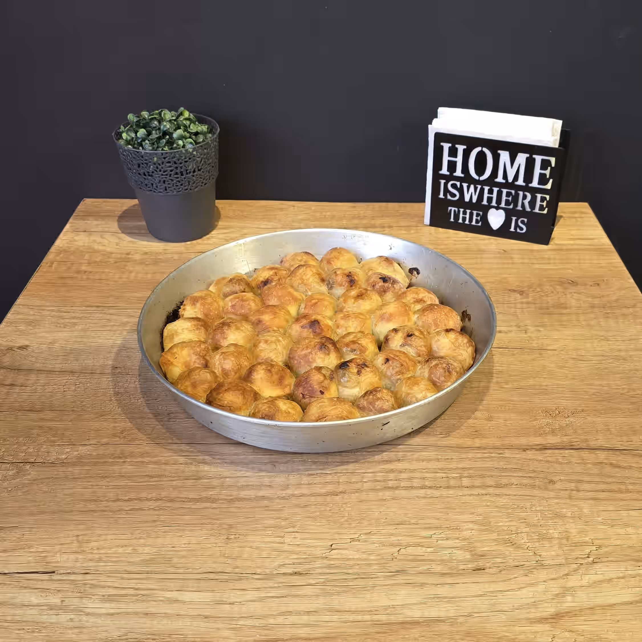 Baking pan filled with golden-brown baked rolls on a wooden table next to a small potted plant and a napkin holder with the text 'HOME IS WHERE THE ♥ IS'.