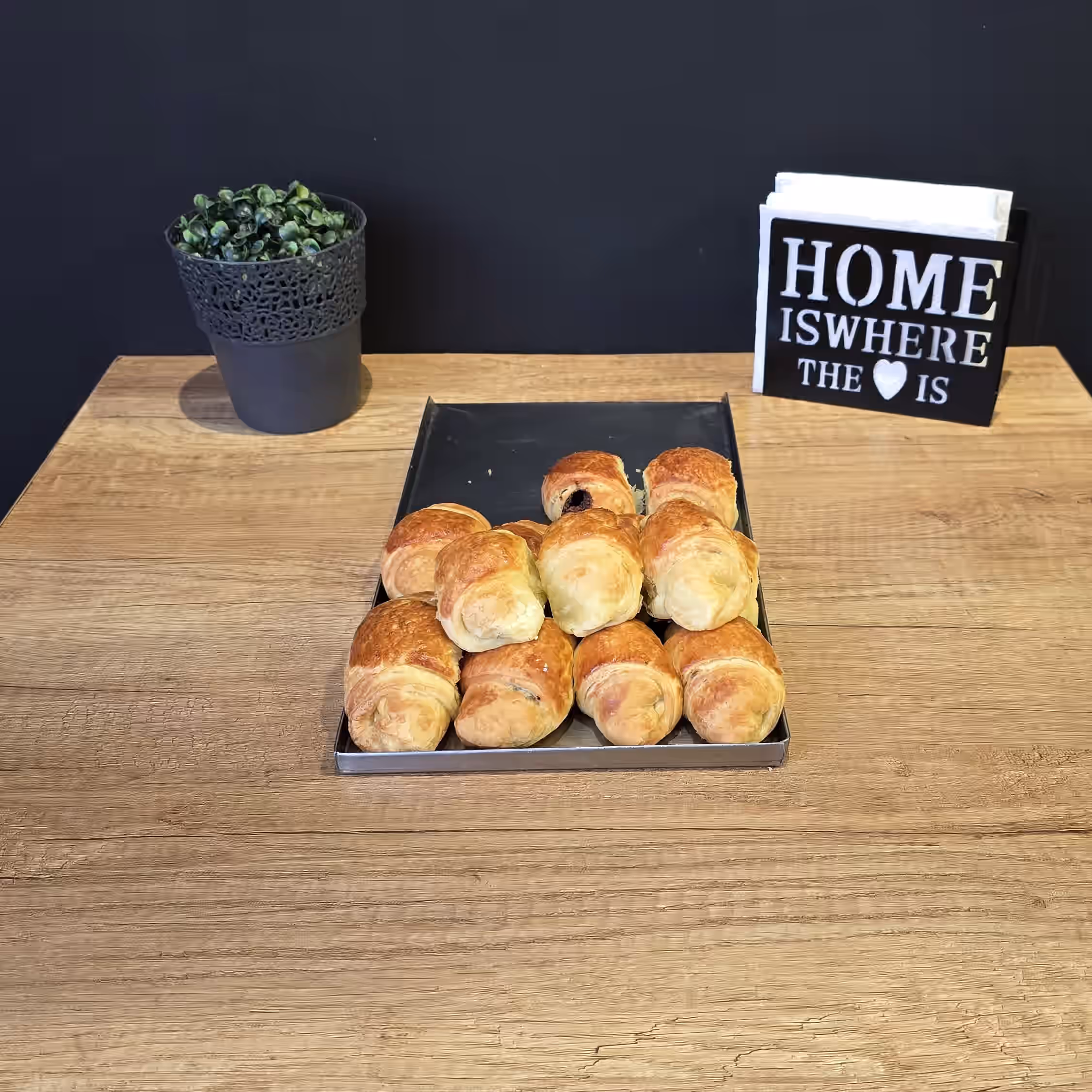 Tray with twelve golden brown croissants on a wooden table with a small potted plant and a sign reading 'HOME IS WHERE THE ♥ IS' in the background.