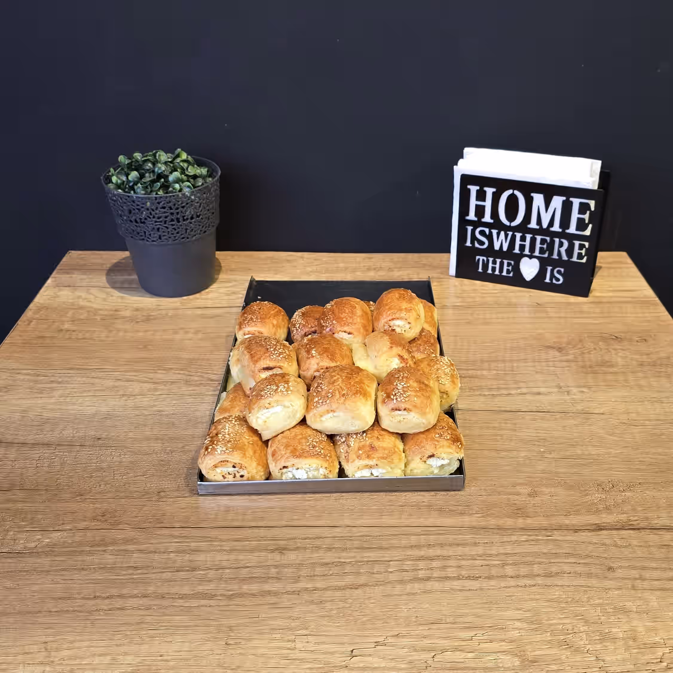 Tray of sesame-topped baked rolls on a wooden table with a potted plant and a black sign that reads 'HOME IS WHERE THE ♥ IS'.