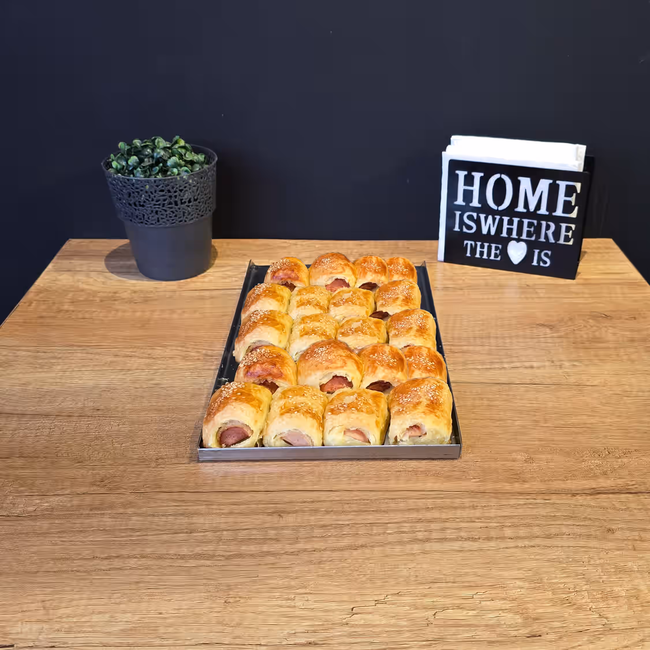 Tray of golden-brown mini sausage rolls sprinkled with sesame seeds on a wooden table, with a small potted plant and a napkin holder reading 'HOME IS WHERE THE ♥ IS' in the background.