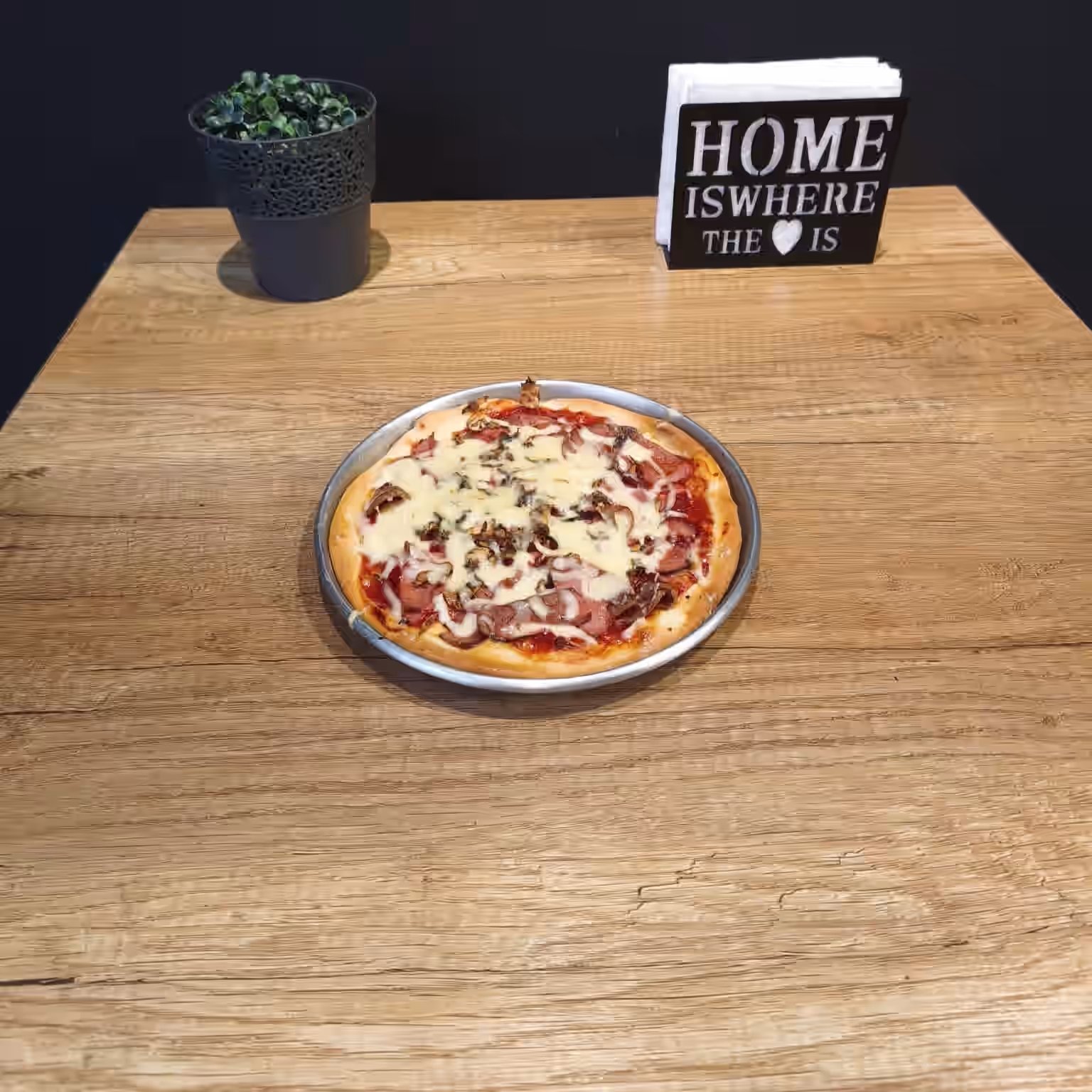 Personal-sized pizza with cheese and toppings on a metal tray on a wooden table with a small plant and a napkin holder reading 'HOME IS WHERE THE ♥ IS'.