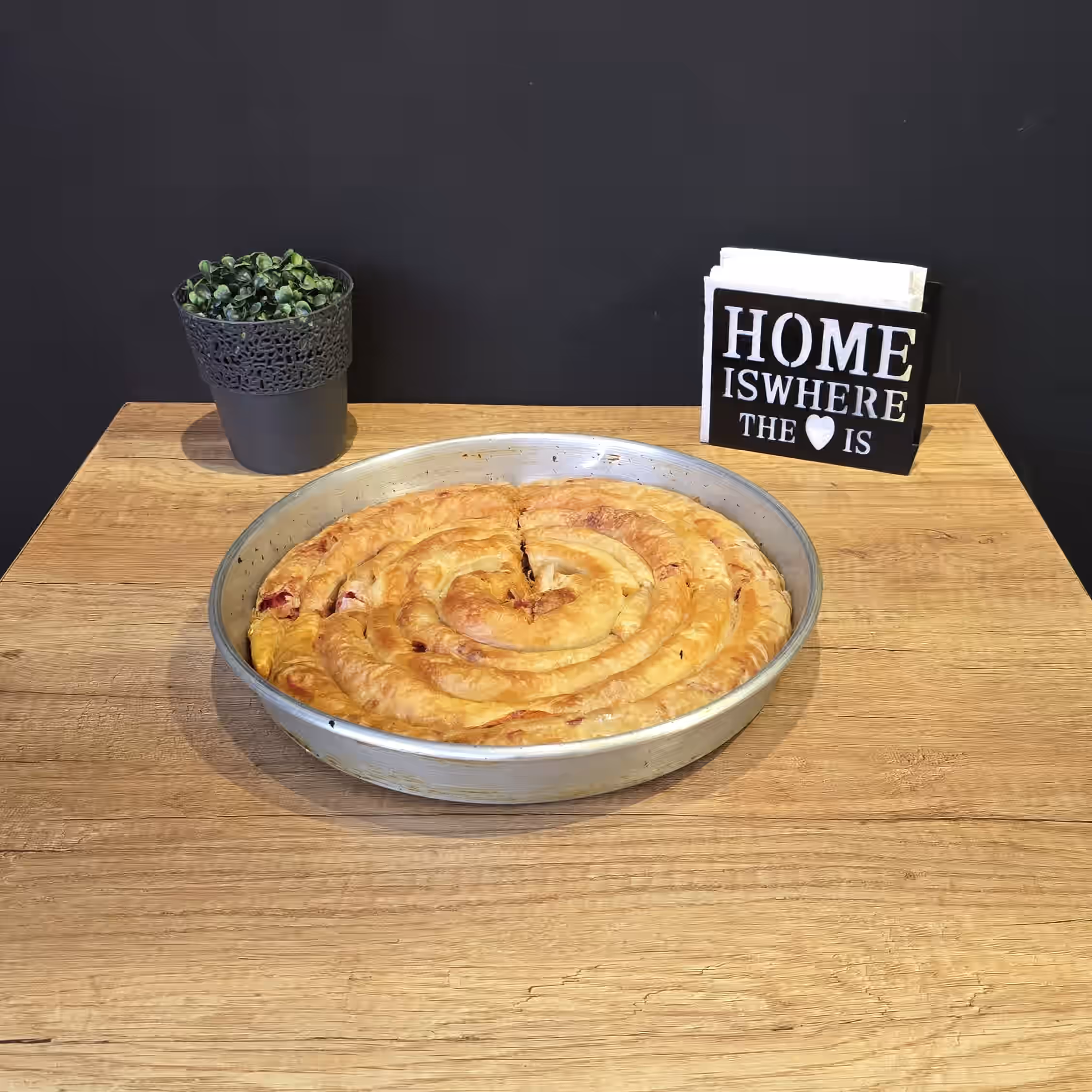 Round spiral-shaped baked pastry in a metal pan on a wooden table with a small potted plant and a black and white wooden sign reading 'HOME IS WHERE THE ♥ IS'.