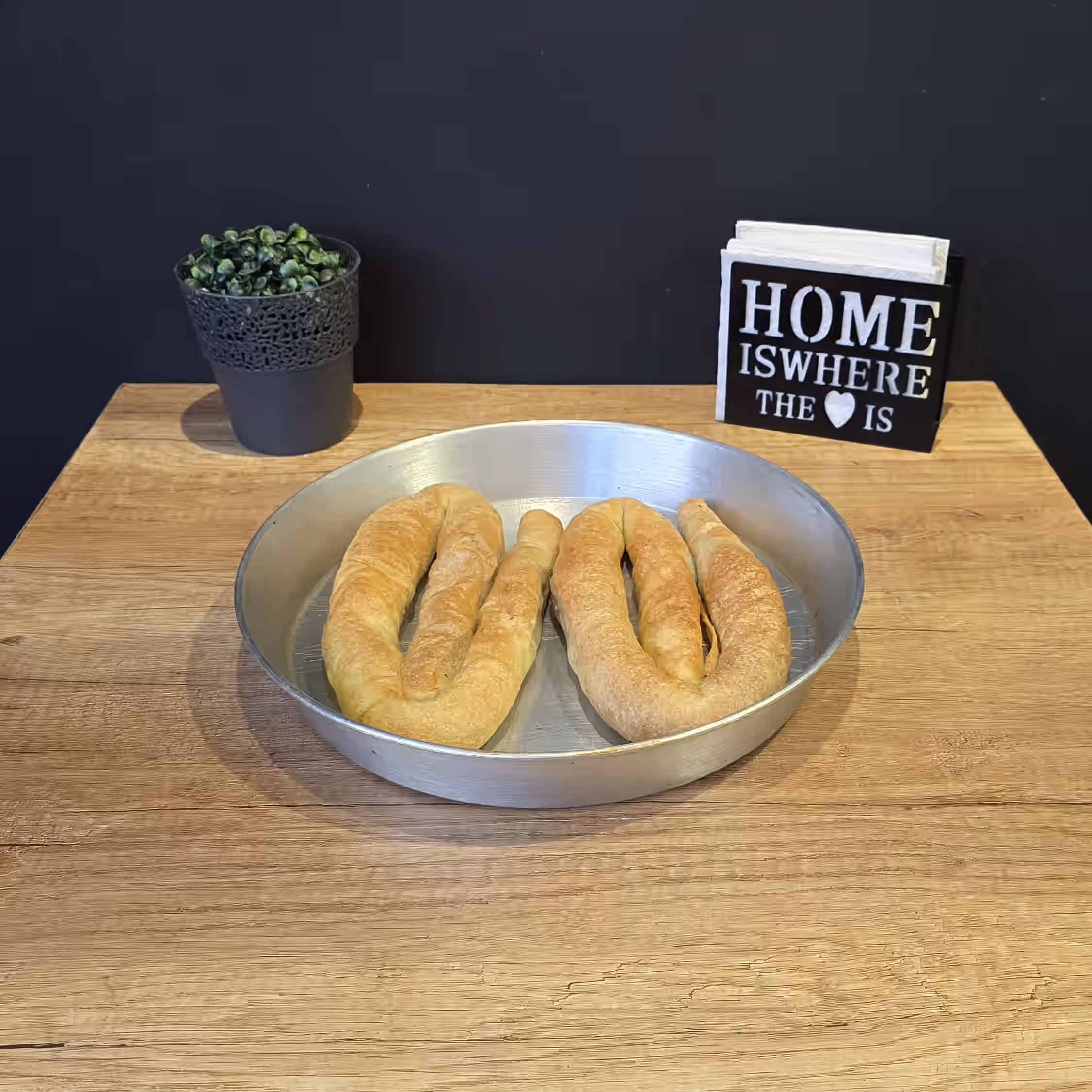 Two crescent-shaped breads in a round metal tray on a wooden table with a small potted plant to the left and a decorative sign reading 'Home is where the heart is' to the right.