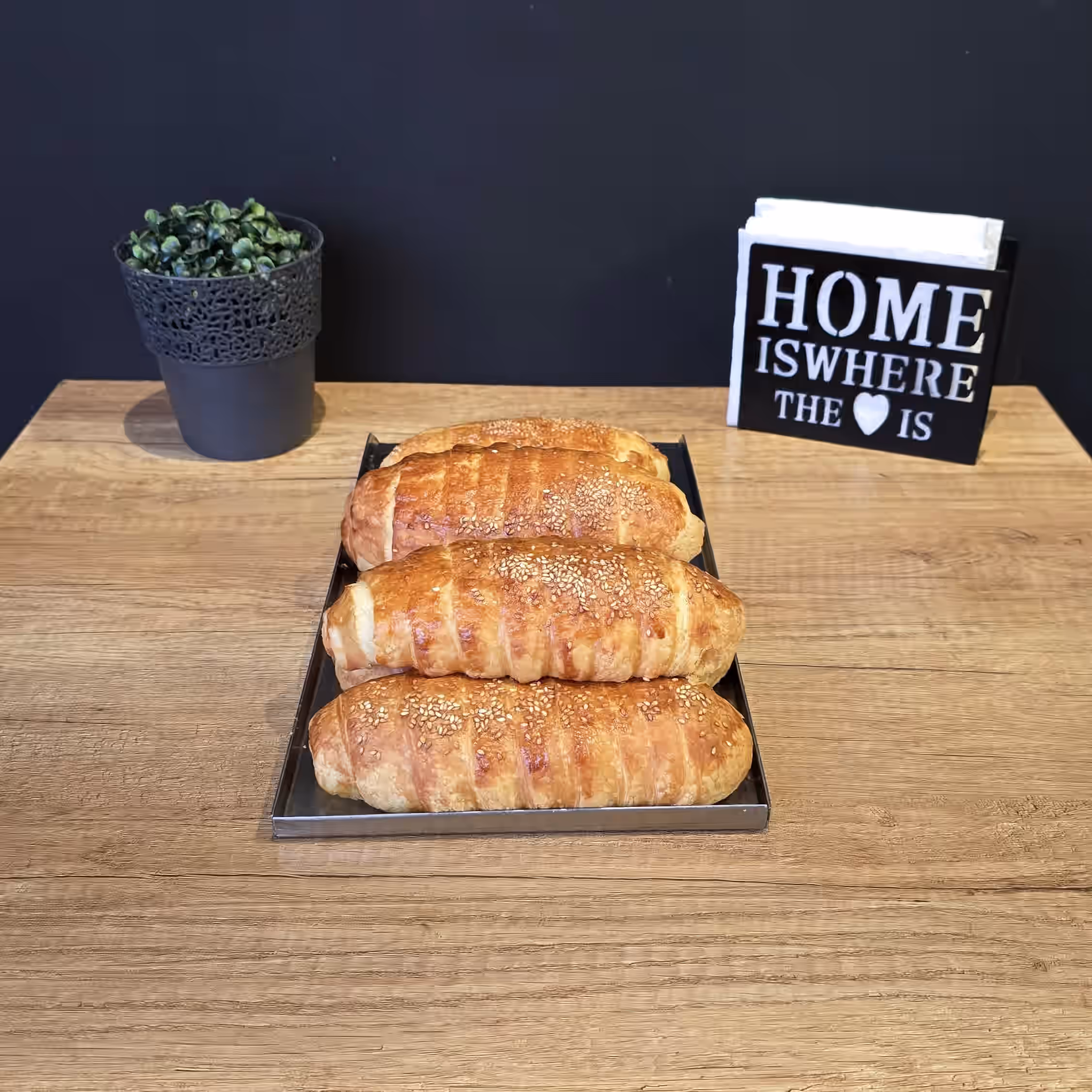 Four golden-brown sesame seed-topped bread rolls on a tray on a wooden table with a small potted plant and a napkin holder with a sign reading 'HOME IS WHERE THE ♥ IS'.