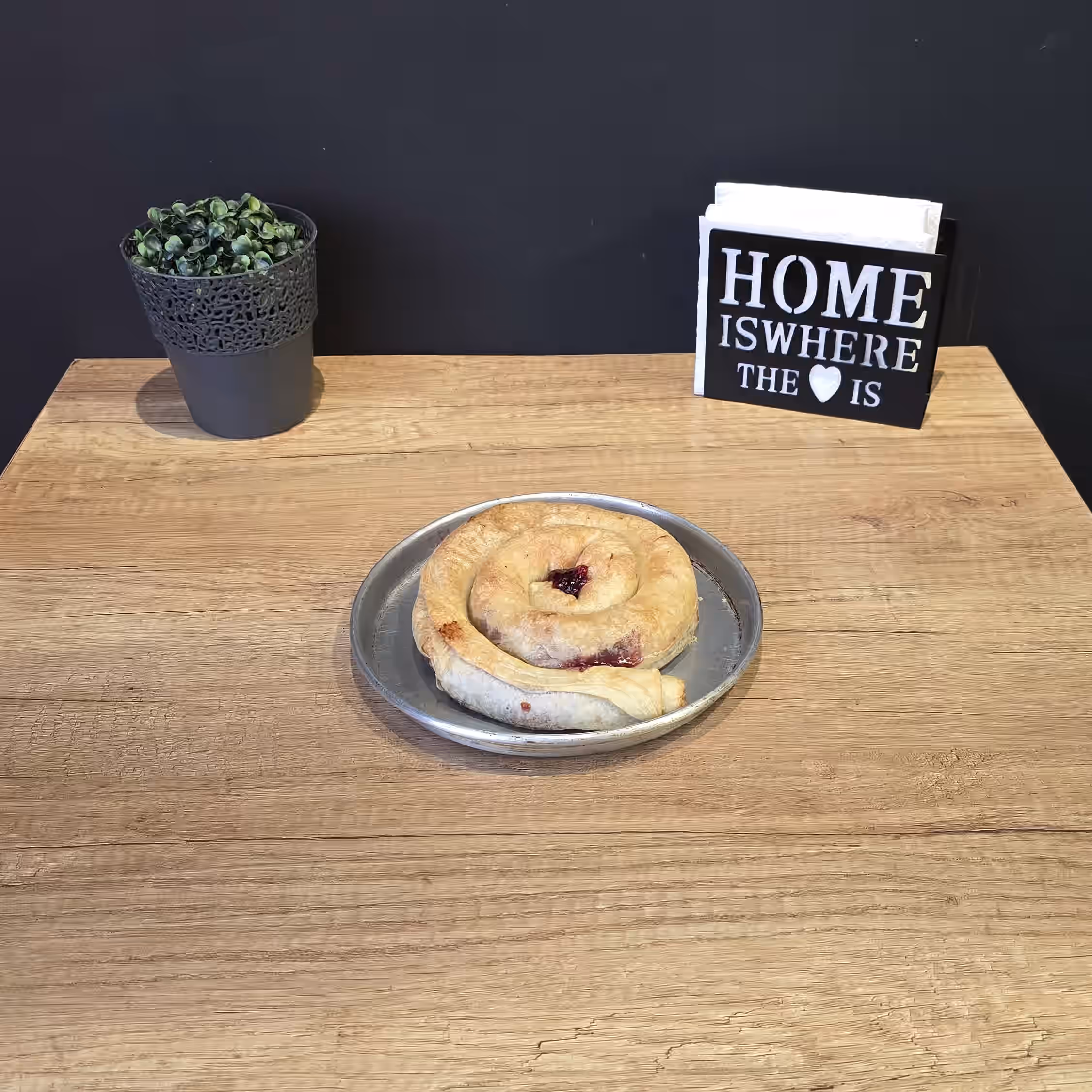 Coiled pastry with berry filling on a metal plate on a wooden table, with a potted plant and a sign reading 'Home is where the heart is' in the background.