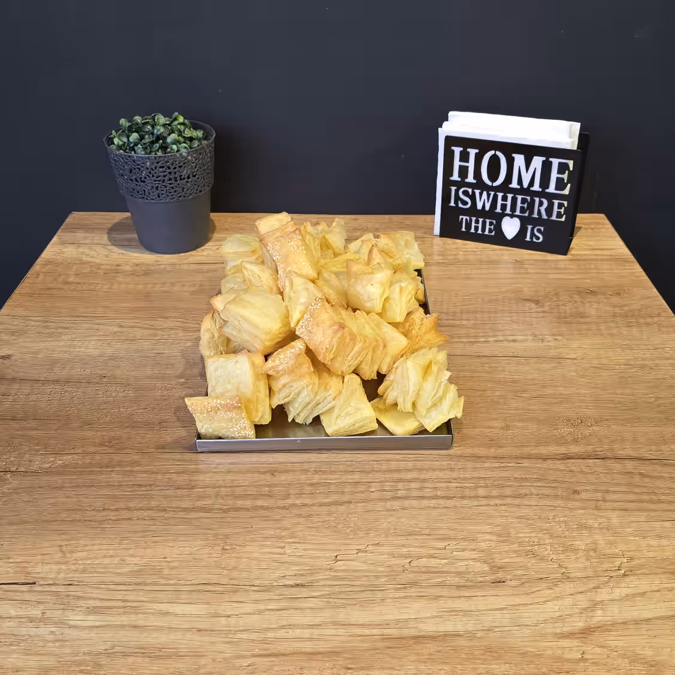 Tray of crispy golden puff pastry squares on a wooden table with a small potted plant and a napkin holder reading 'HOME IS WHERE THE ♥ IS' against a dark background.